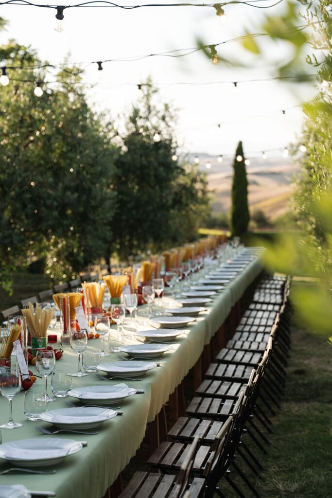 Lange gedeckte Tafel mit grüner Tischwäsche, Tellern und italienischer Dekoration unter Lichterketten im Olivenhain.