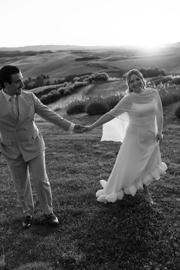 Schwarz-Weiß-Aufnahme von Sophie und Erik, die sich an den Händen halten und gemeinsam durch die hügelige Landschaft der Toskana gehen.