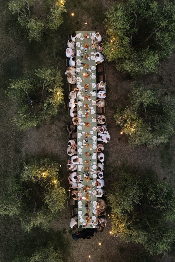 Hochzeitsgäste stoßen an einer langen gedeckten Tafel unter Lichterketten bei einer italienischen Pizza-Party in der Toskana an.