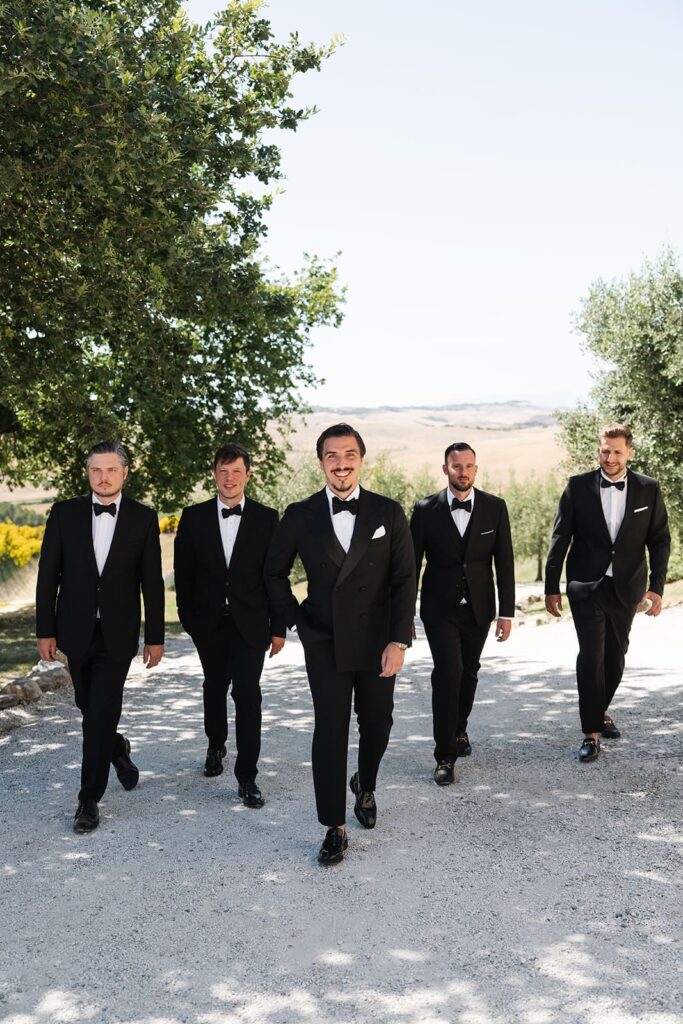 Bräutigam Erik und seine Groomsmen in schwarzen Anzügen spazieren gemeinsam durch den Garten einer Hochzeitslocation in der Toskana.