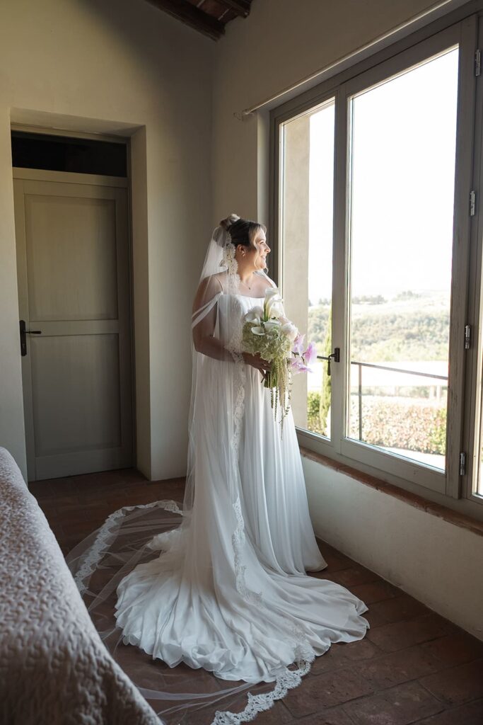 Braut Sophie steht in ihrem weißen Hochzeitskleid mit langem Schleier und Brautstrauß vor einem großen Fenster mit Blick in die Toskana.