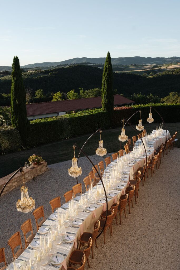 Lange festlich gedeckte Hochzeitstafel mit hängenden Kronleuchtern und Blick auf die toskanische Landschaft im Abendlicht.
