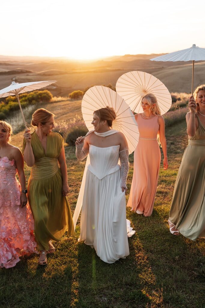 Braut Sophie steht mit ihren Brautjungfern in rosa und grünen Kleidern im warmen Abendlicht, während sie dekorative Fächer halten.