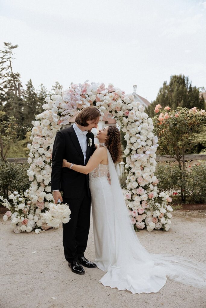 Sali und Szymon küssen sich vor einem opulenten Blumenbogen mit weißen und rosafarbenen Blüten bei ihrer freien Trauung.