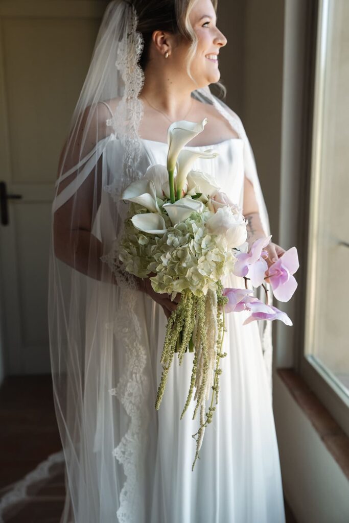 Braut Sophie in ihrem eleganten Hochzeitskleid mit Brautstrauß am Fenster einer Hochzeitslocation in der Toskana.