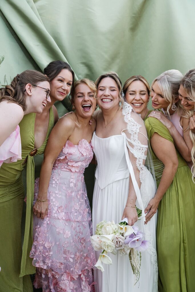 Braut Sophie lacht gemeinsam mit ihren Brautjungfern in rosa und grünen Kleidern vor einem grünen Stoffhintergrund bei ihrer Hochzeit in der Toskana.