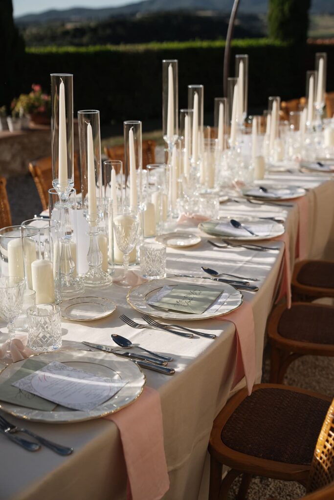 Nahaufnahme einer elegant gedeckten Hochzeitstafel mit weißen Tellern, Gläsern, hohen Glaszylindern und stilvoller Tischdekoration.