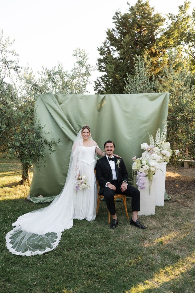 Brautpaar Sophie und Erik vor einem salbeigrünen Stoffhintergrund mit eleganten Blumenarrangements im Garten ihrer Hochzeitslocation in Volterra.